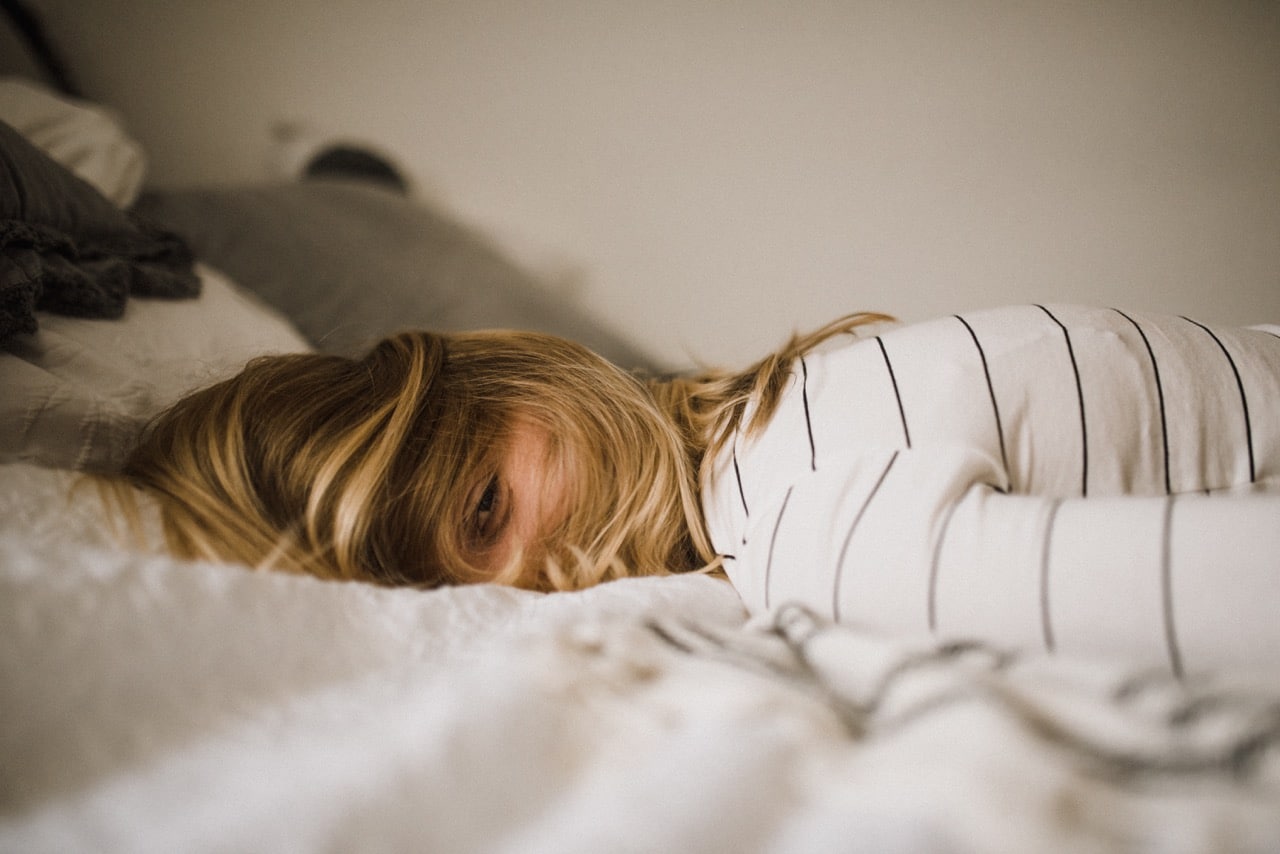 Das Bild zeigt eine müde Frau bäuchlings auf einem Bett liegend mit einem Auge geöffnet.